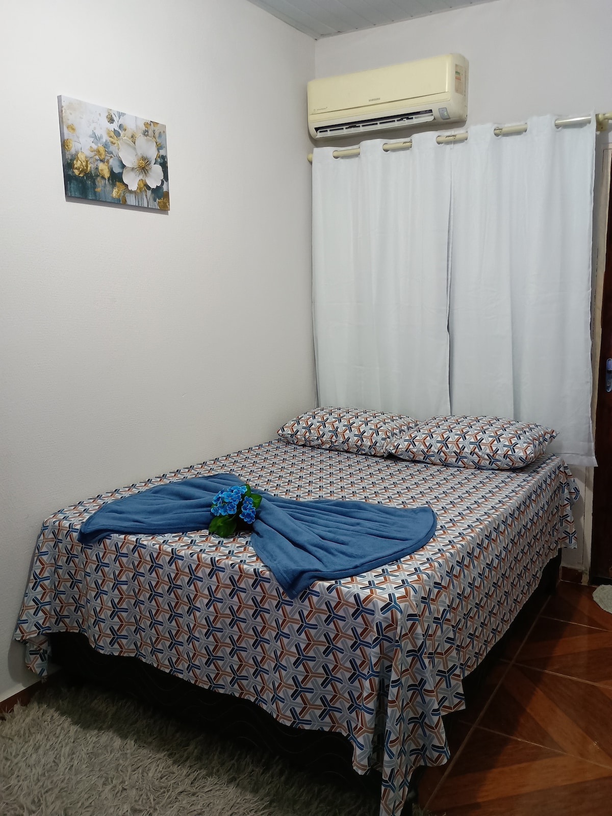 A cozy sleeping area features a spacious bed covered in a patterned blanket. Two pillows are neatly arranged against the headboard. A small floral wall art is displayed above the bed, and light enters through sheer white curtains, complemented by an air conditioning unit on the wall.