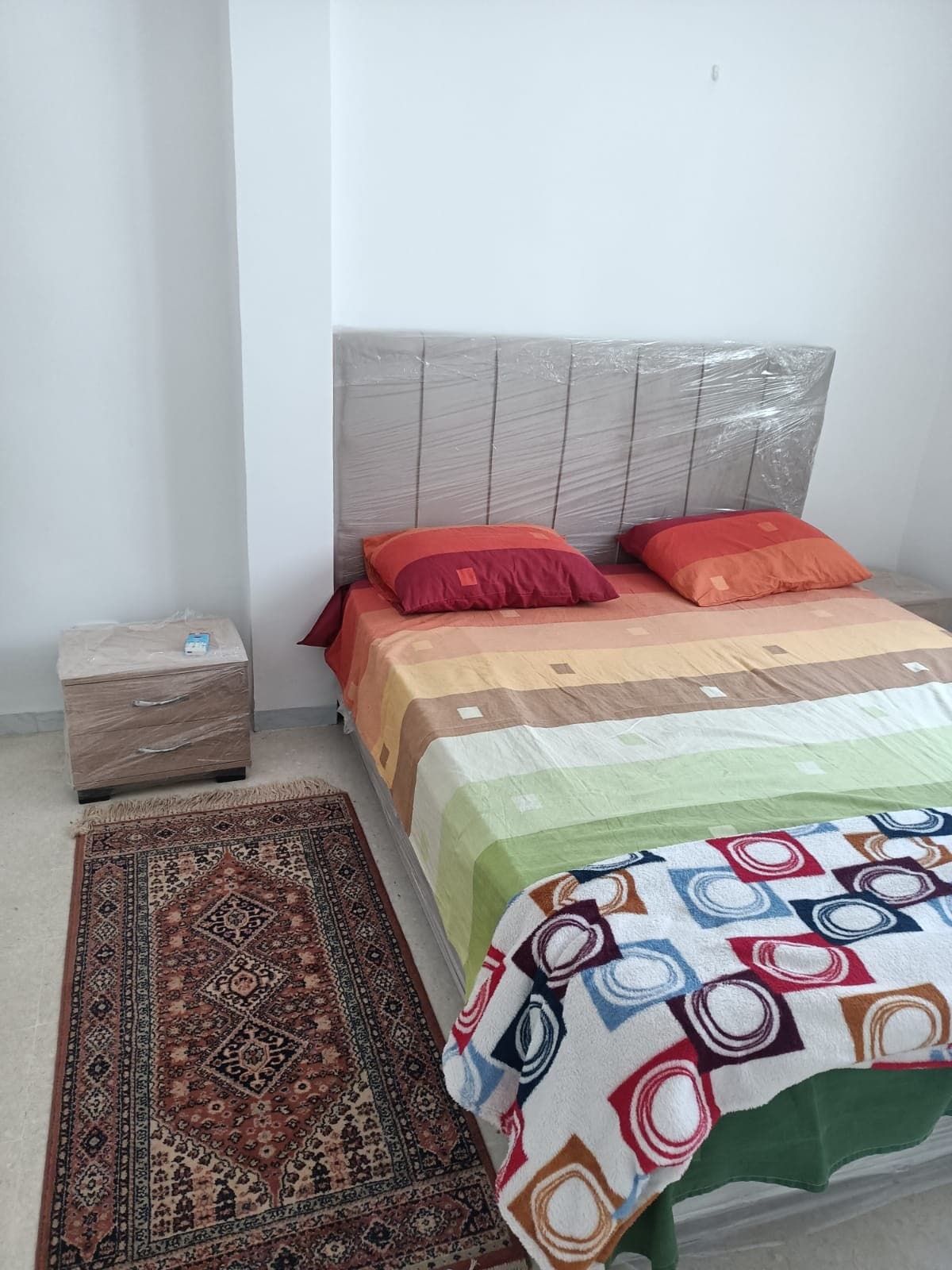 A light-filled sleeping area features a spacious bed with a neutral headboard, adorned with colorful bedding in shades of orange and green. A bedside table holds a small blue item, and a patterned rug adds texture to the floor.