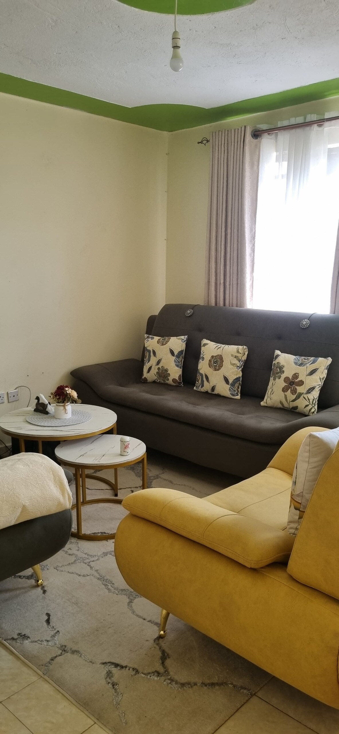 A comfortable living area features a dark gray sofa with floral cushions and a yellow armchair. A round coffee table with a decorative item occupies the center. Natural light enters through a window, complemented by soft curtains.