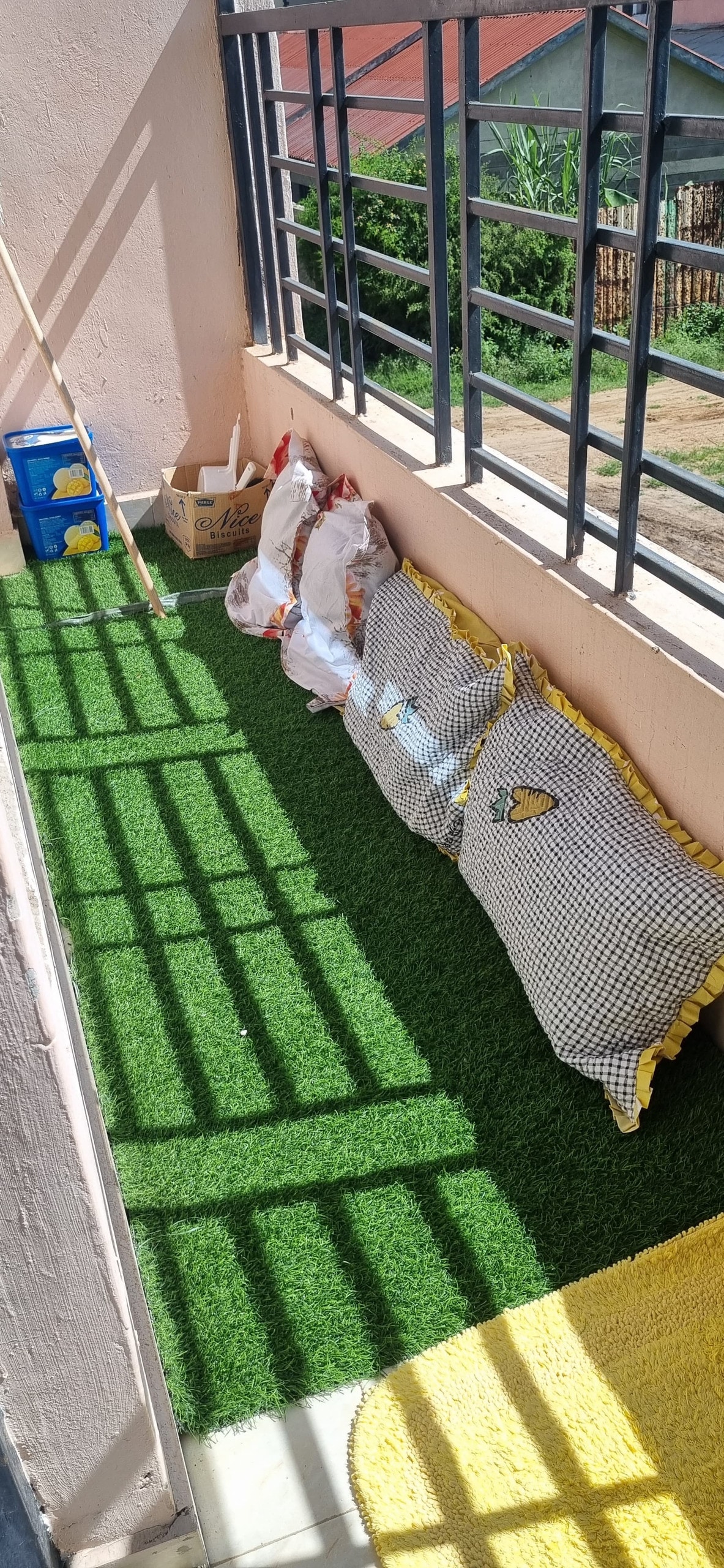 A small balcony area features a green artificial grass surface with cushions against the wall for seating. Shadows from the railing create patterns on the ground, while a few bags are arranged nearby. Complementary decor elements add an inviting touch to the space.