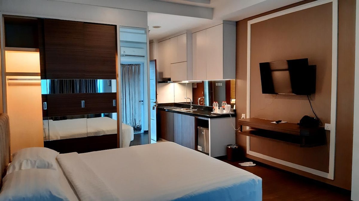 A modern hotel room features a spacious bed against a sleek wall unit with mirrored elements. A kitchenette is visible, equipped with essential appliances, while a wall-mounted television sits opposite the bed. Natural light enters through a nearby window, enhancing the room's inviting ambiance.