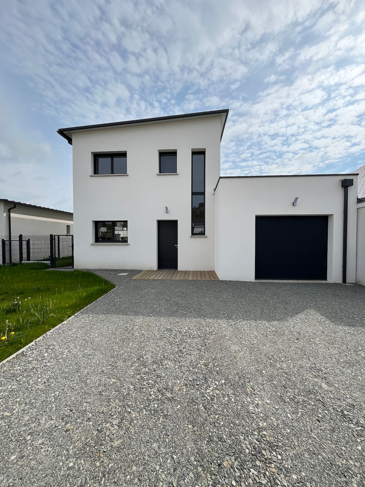 The front of the modern house is shown, displaying large windows and a sleek entrance door. A wooden deck leads to the doorway, while a neatly gravel-lined driveway provides access to the attached garage. The surrounding green grass complements the contemporary design.