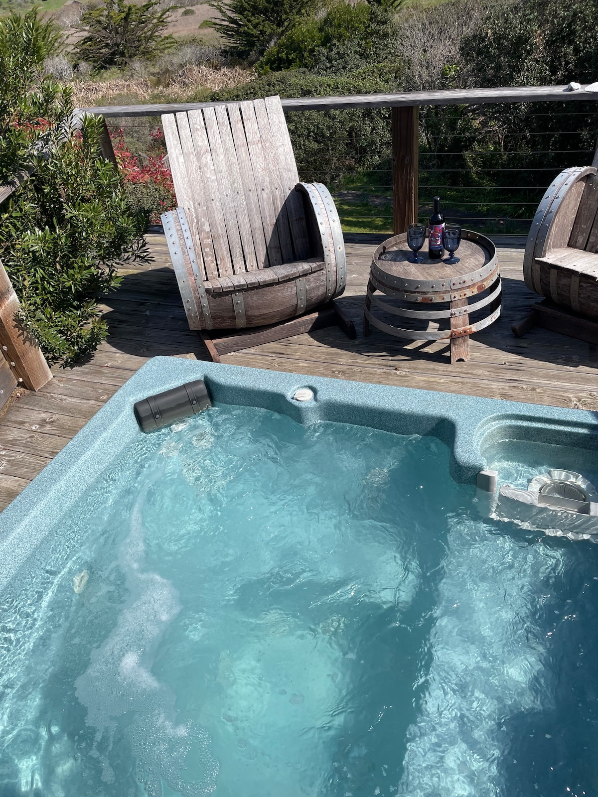 A private hot tub is nestled on a wooden deck, featuring clear, inviting water. Two rustic chairs made from reclaimed wood sit nearby, accompanied by a small barrel table topped with two wine glasses. Lush greenery frames the space, enhancing the secluded feel.