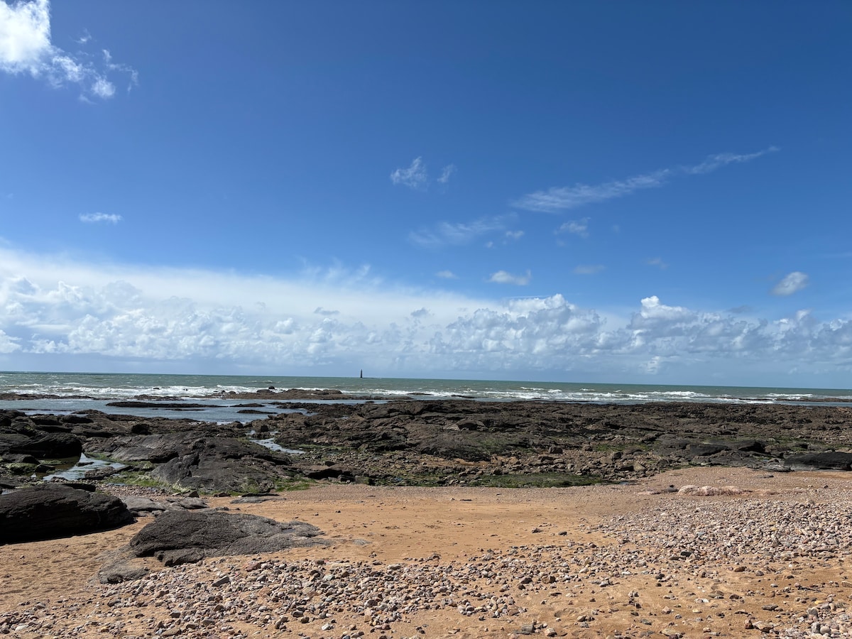 A view of the rocky beach stretches out towards the ocean, with gentle waves meeting the shore under a clear blue sky. Scattered boulders and small pebbles cover the sandy area, while a distant lighthouse stands sentinel on the horizon, framed by clouds.