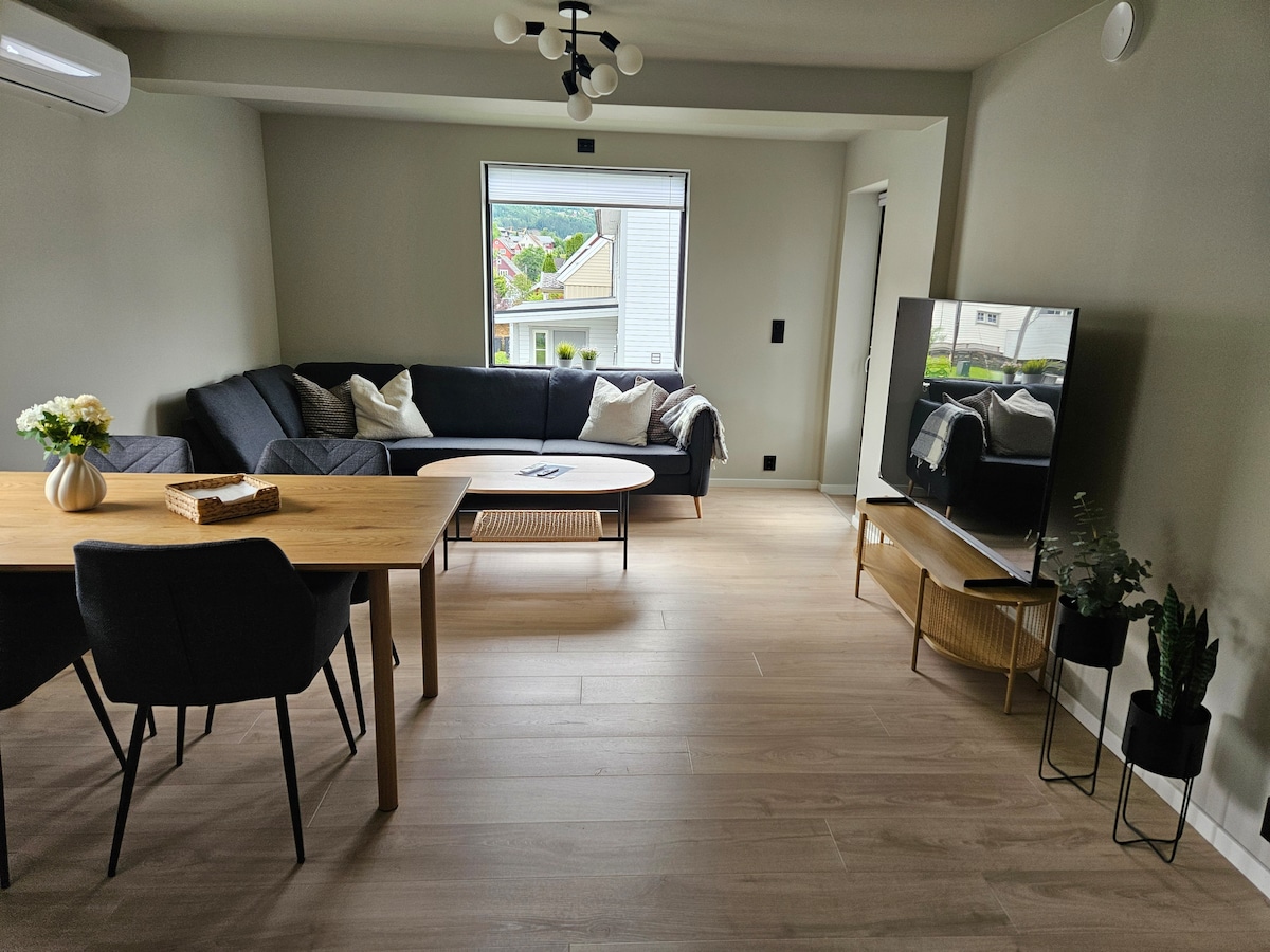 The living area features a dark sectional sofa paired with a round coffee table. A wooden dining table with chairs is present, and a television sits on a stand beside a decorative plant. Natural light enters through the window, enhancing the soft color palette.