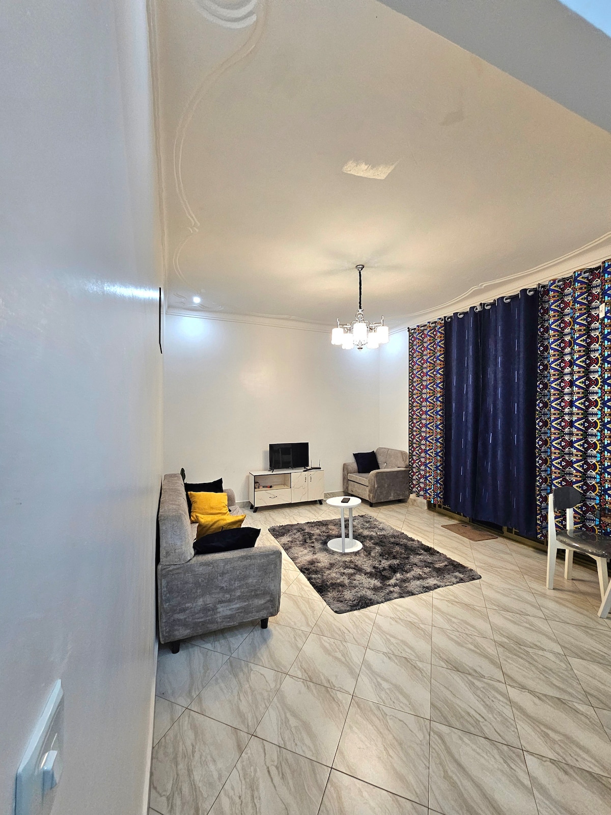 A well-lit living room features a cozy gray sofa accented with a yellow throw pillow. A television stands on a white cabinet, with a round coffee table positioned on a plush area rug. Two patterned curtains frame a window, adding a pop of color to the space.