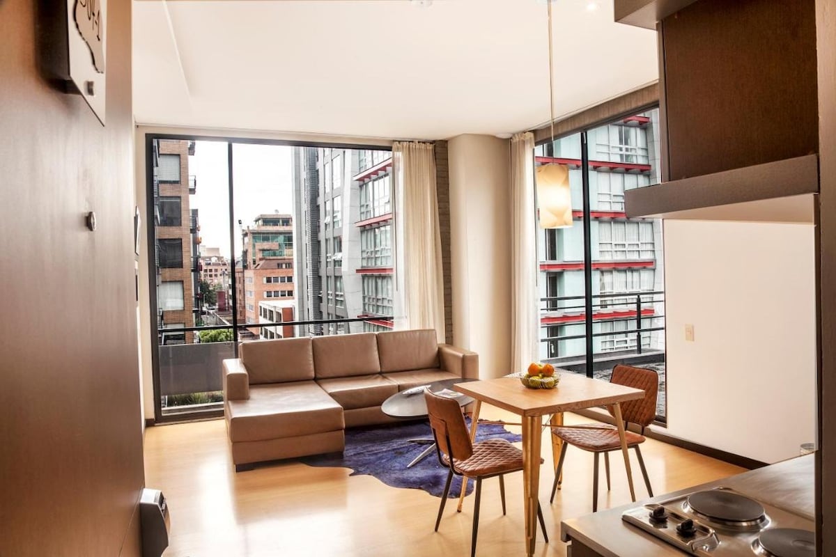 A spacious living area is captured, featuring a comfortable couch and a small dining table with two chairs. Large windows allow natural light to brighten the room, while city views create a lively atmosphere. A fruit bowl sits on the table, adding a touch of warmth.