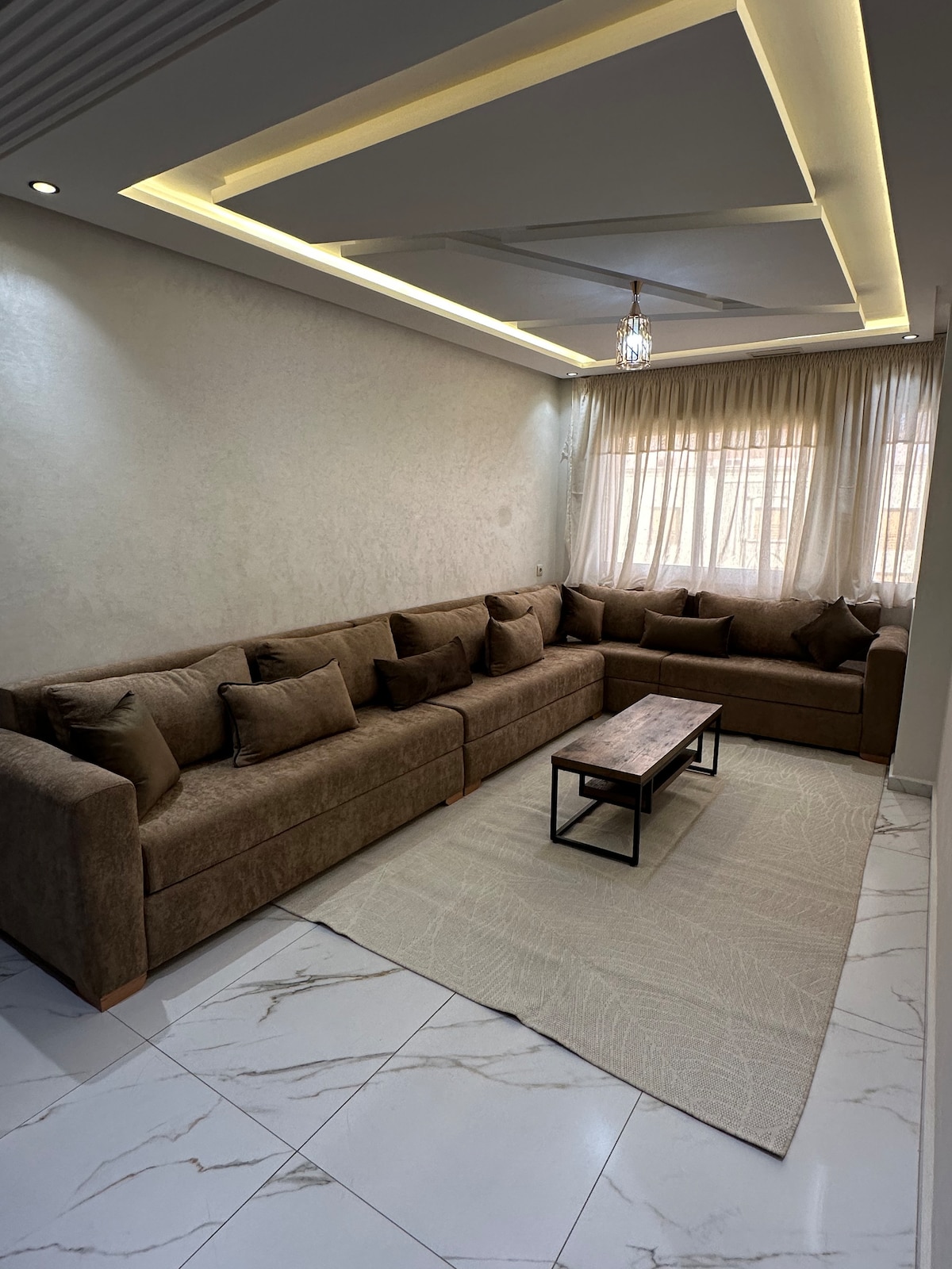 A spacious living area features a large, L-shaped brown sofa with multiple cushions, positioned around a rectangular wooden coffee table. Light gray curtains frame the window, allowing natural light to illuminate the room. A neutral-toned area rug lies beneath the seating arrangement.