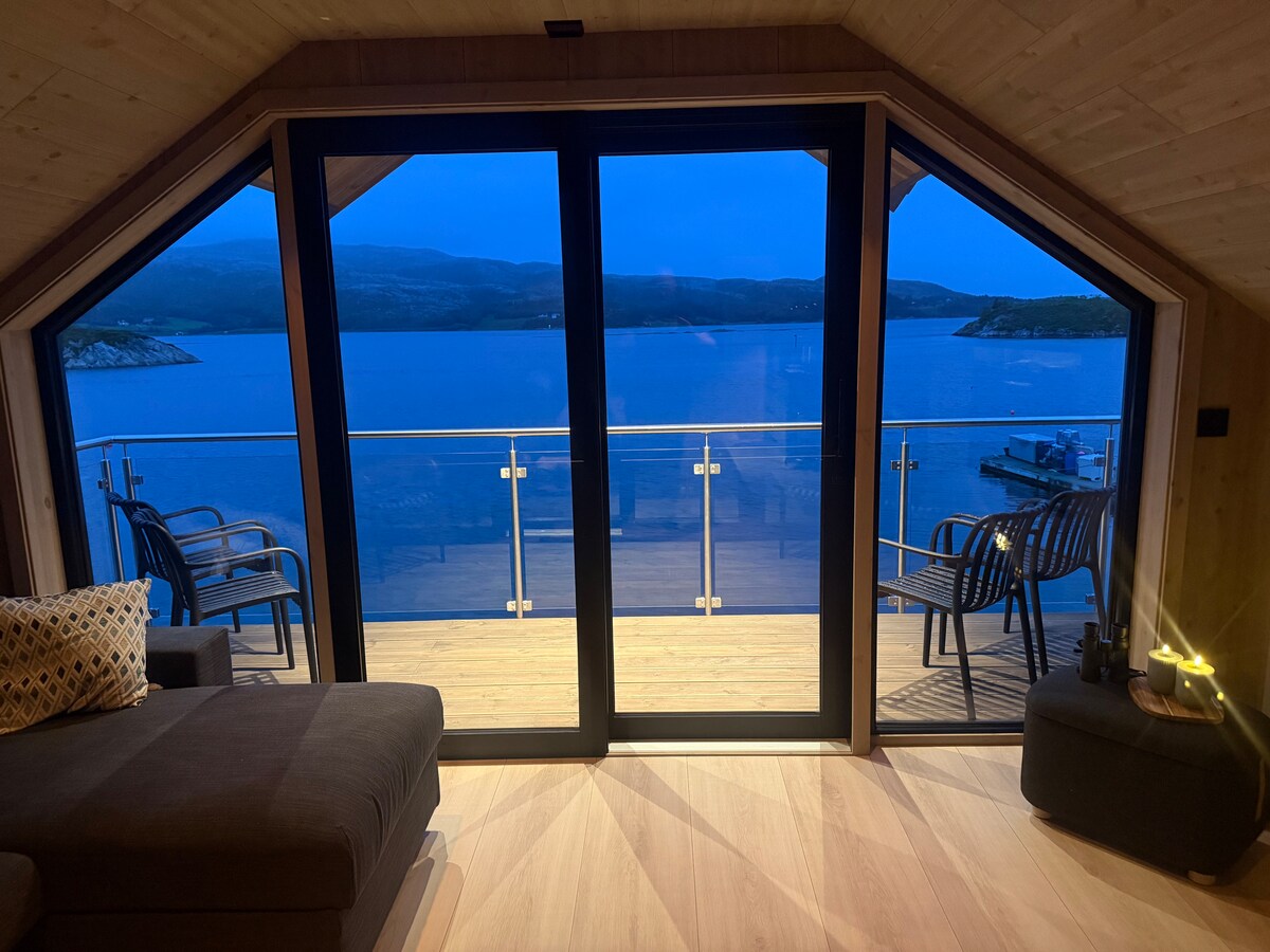 A spacious living area features large glass doors opening onto a private balcony overlooking the water. Black chairs are positioned on the balcony, offering a place to relax while enjoying views of the calm sea and distant mountains. Soft evening light creates a serene atmosphere.