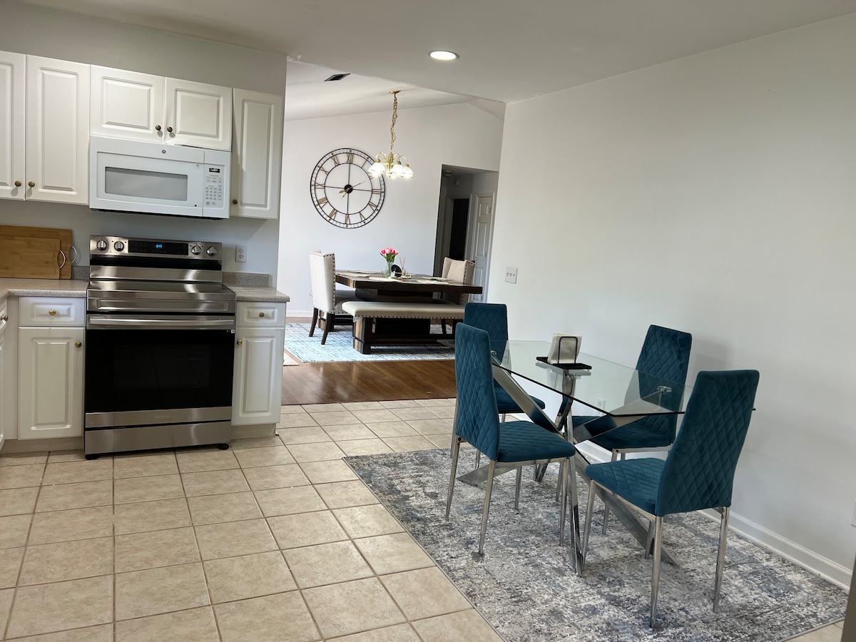 A clean and modern kitchen area features stainless steel appliances and white cabinetry. A glass dining table is surrounded by four blue upholstered chairs on a decorative rug, offering a comfortable space for meals. Natural light brightens the room from a nearby window.