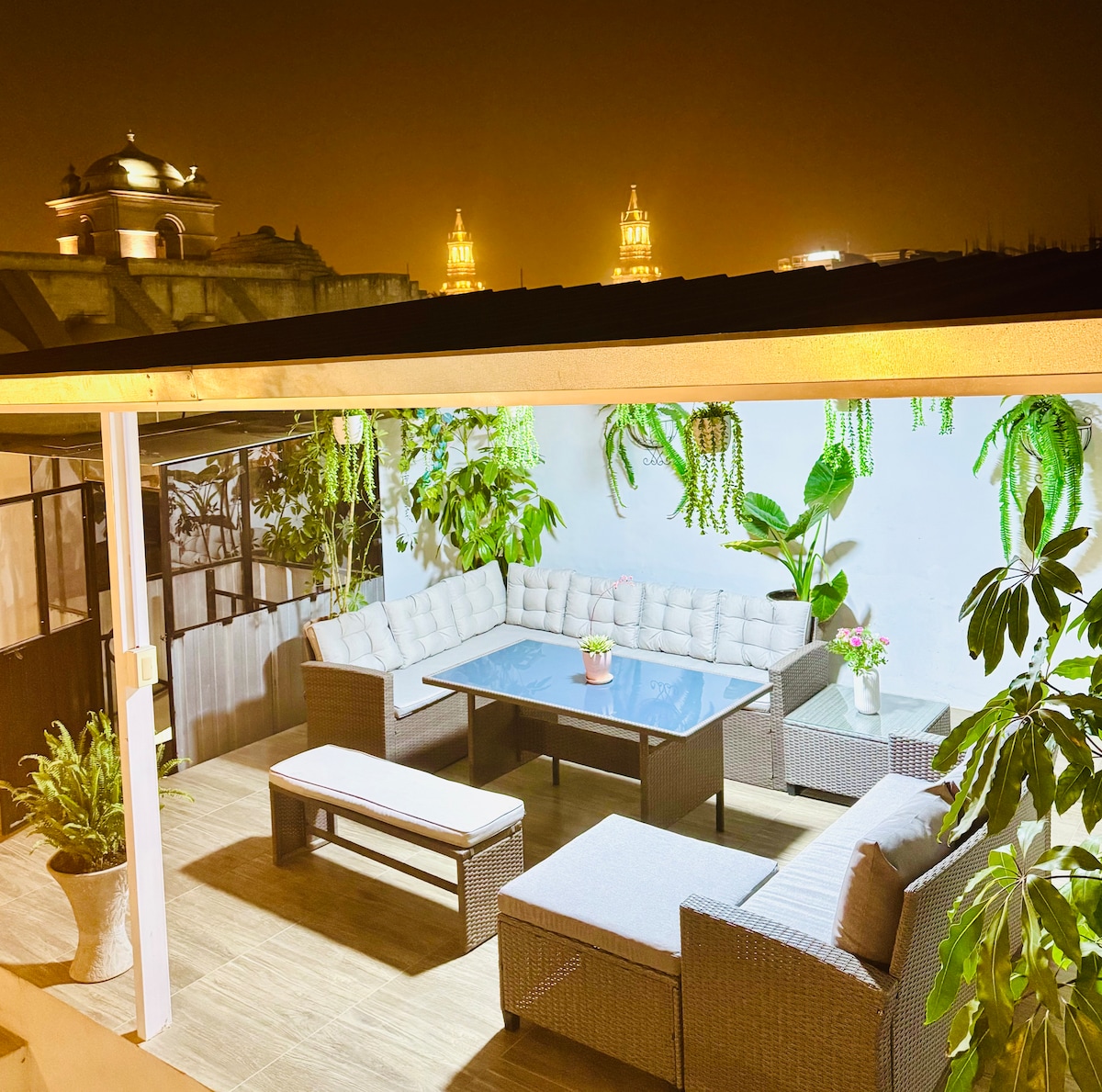 A spacious terrace is furnished with comfortable seating and a central table surrounded by greenery. Soft lighting highlights the outdoor space, with historic towers visible in the distance, creating a relaxed atmosphere for gatherings.