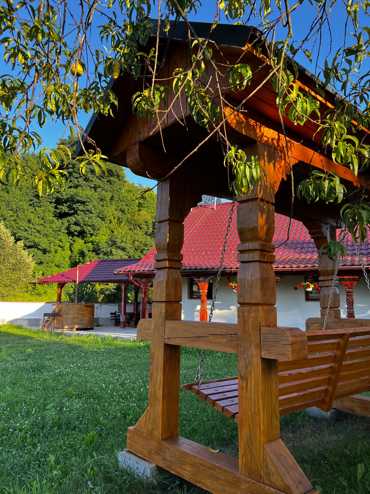 A traditional wooden swing can be seen in the foreground, surrounded by lush greenery. In the background, a charming house with a red roof is partially visible, alongside a patio area that hints at outdoor leisure.