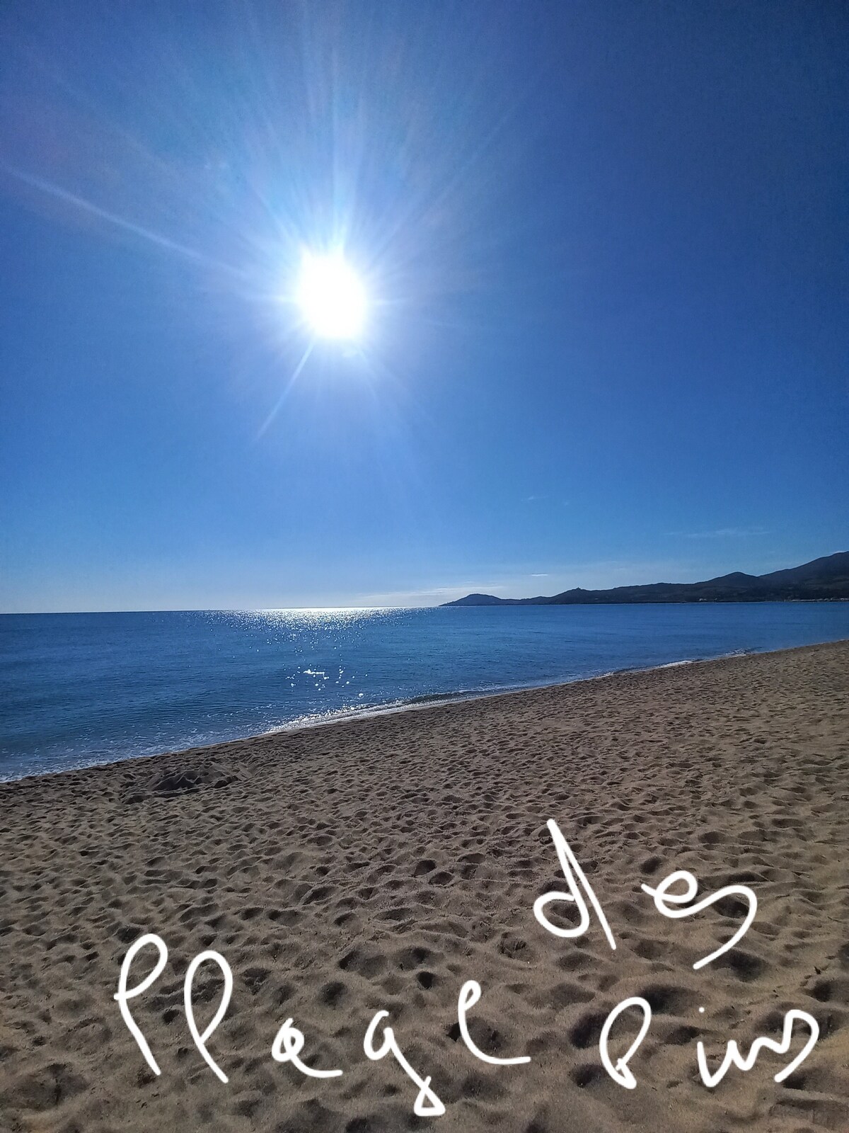 A sandy beach is depicted under a bright sun, with clear skies and a serene ocean view. The gentle waves reflect sunlight, enhancing the tranquil atmosphere. Rolling hills are visible in the distance, framing this coastal landscape.