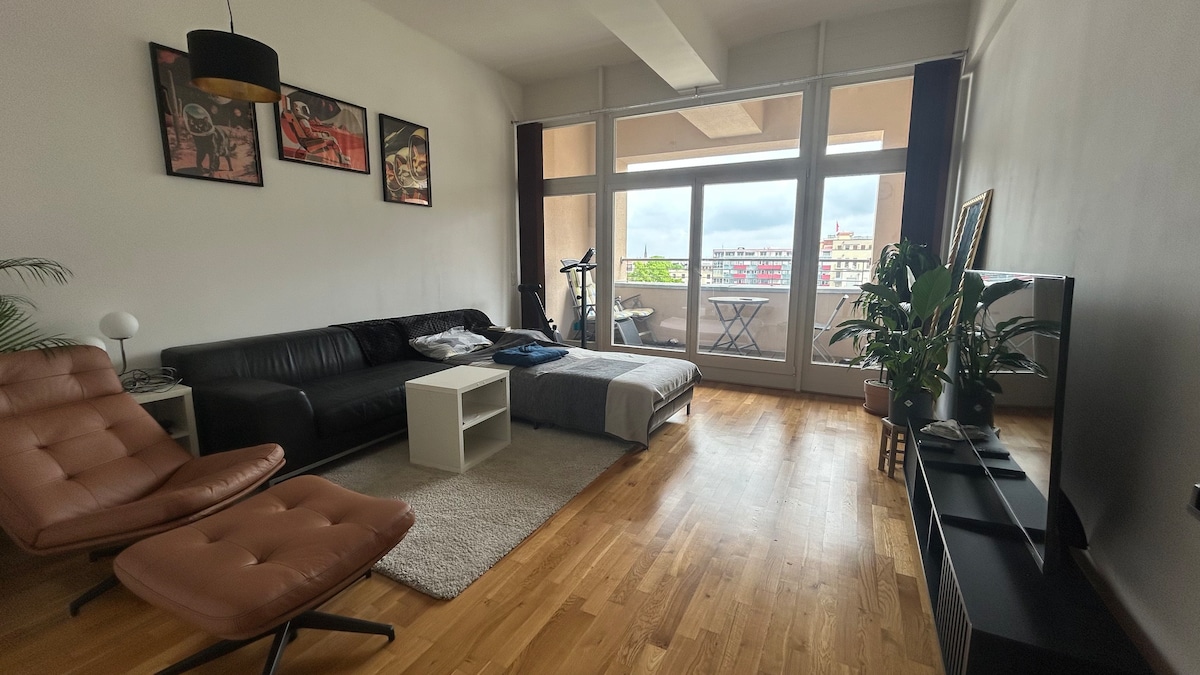 A modern living area is showcased with a black sofa and a brown lounge chair. A light-colored coffee table sits center stage on a plush rug. Large windows provide natural light, revealing a view of the balcony and outdoor seating, with potted plants enhancing the space.