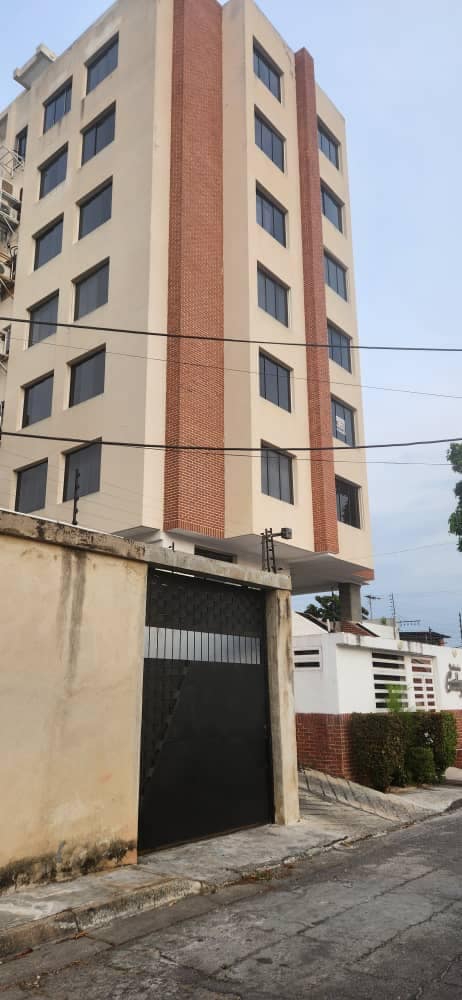 A modern six-story building features a combination of brick and cream-colored facades. Large, evenly spaced windows provide natural light. The entrance is marked by a secure, gated access, and neatly maintained greenery lines the perimeter.