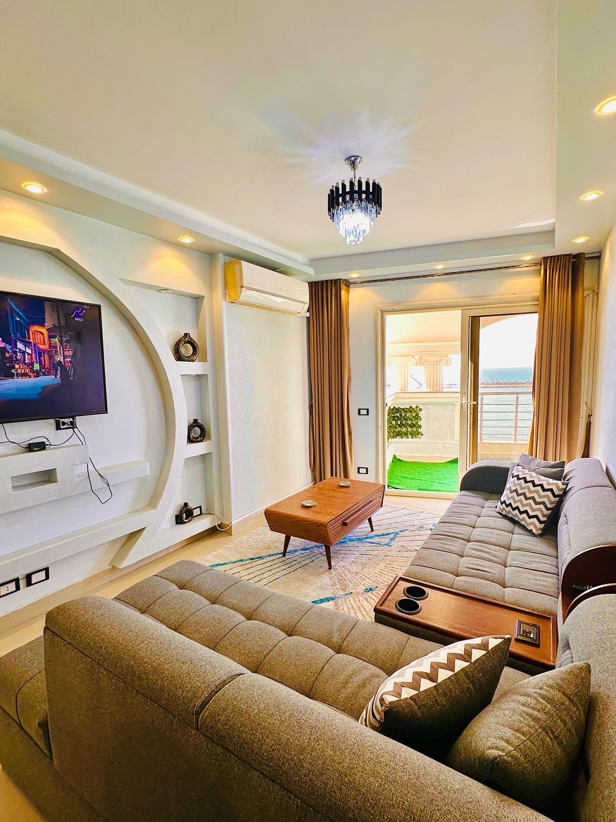 A living area is presented with a comfortable sofa, accompanied by a coffee table. A wall-mounted television is visible, and a large window offers a view of the outdoor space. Natural light is drawn in through the curtains, highlighting the modern decor.