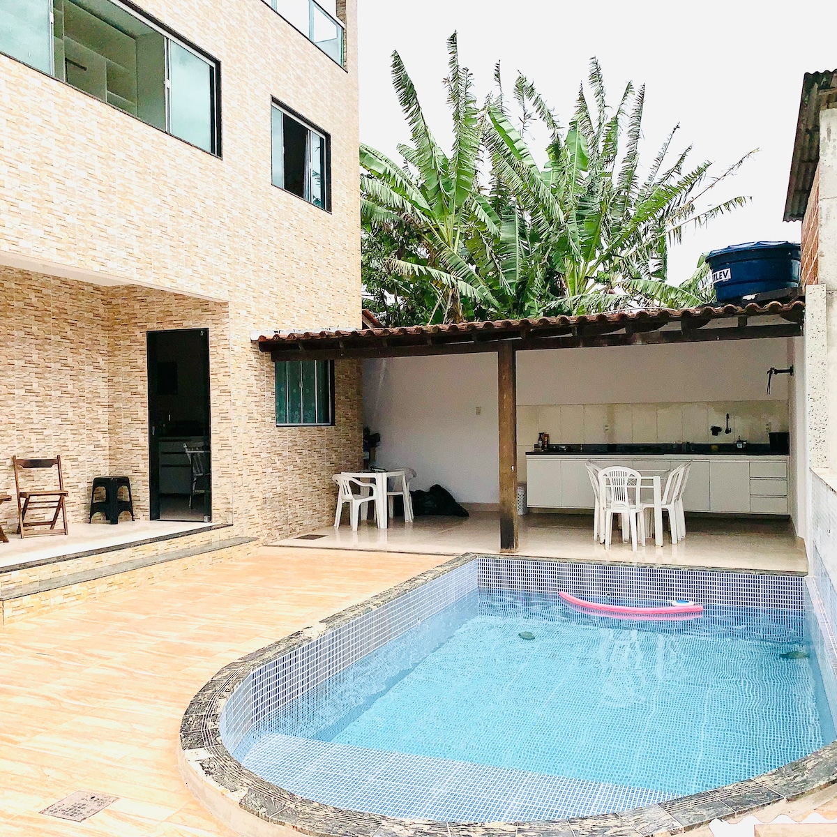 A private outdoor area features a swimming pool bordered by tiles, complemented by a shaded space containing a table and chairs. Lush green plants provide a natural backdrop, while a partially covered area includes a kitchen space. The inviting setting offers ample room for relaxation.