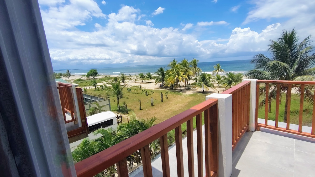 A balcony view showcases the expansive beach and ocean, framed by swaying palm trees and a manicured lawn. The blend of blue skies and scattered clouds creates a peaceful backdrop, while the calm sea stretches into the distance.