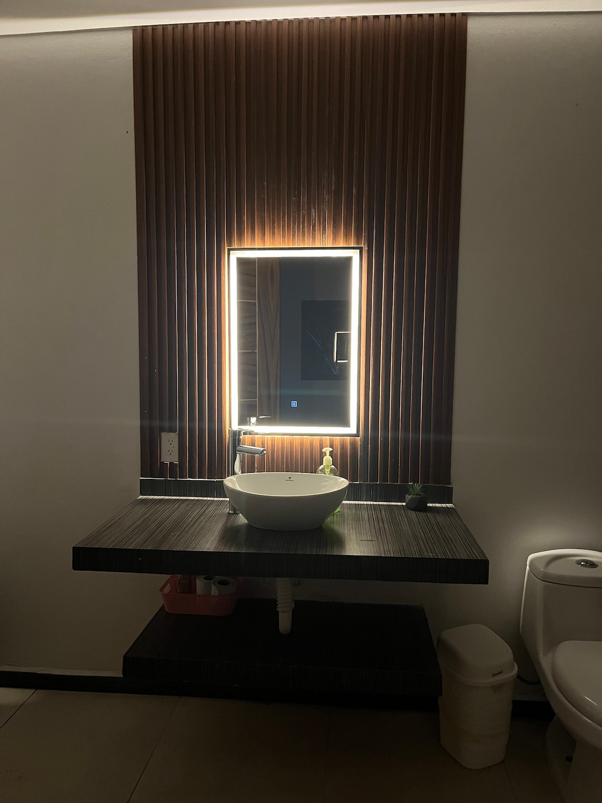 A modern bathroom is presented with a stylish countertop and a round sink. A backlit mirror is mounted above the sink, creating a soft glow. Wooden paneling adds warmth to the wall, while a toilet and waste bin are positioned discreetly in the corner.