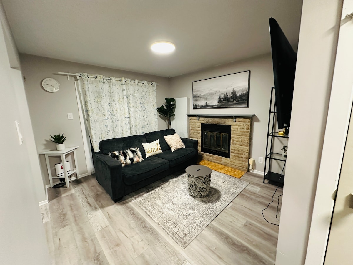 The living area features a dark sofa adorned with cushions, positioned near a stone fireplace. A circular coffee table rests on a patterned rug, while a decorative plant and a wall clock are placed in the corners. Light enters through sheer curtains, enhancing the cozy atmosphere.