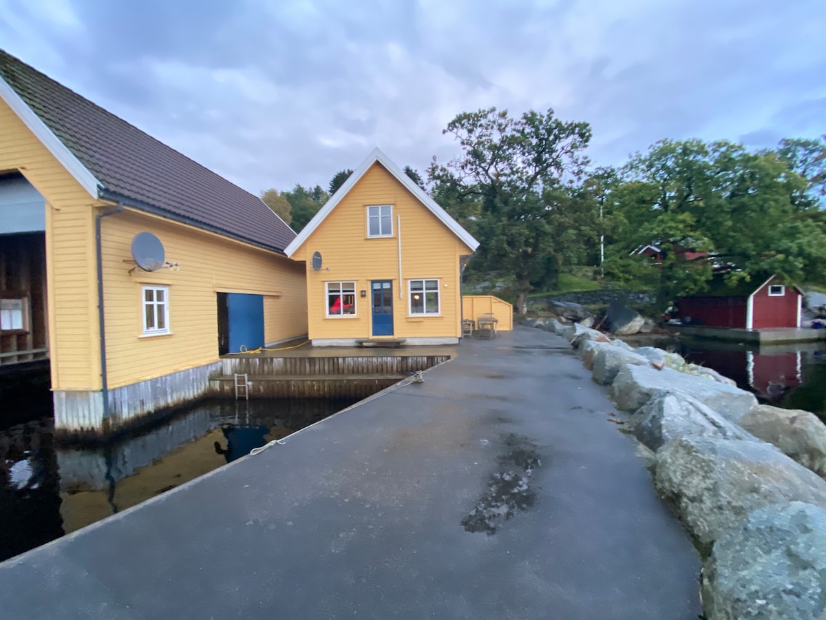 A charming yellow cabin is situated near a smooth stone pier, surrounded by water and greenery. The structure has a blue door and several windows. Outdoor seating is provided on the patio, overlooking the calm waters and the wooded landscape beyond.