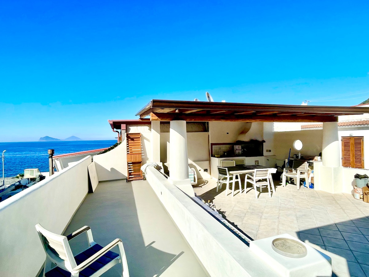 An extensive terrace features outdoor furniture, including sun loungers and a dining area. A barbecue area is visible under a shaded structure, with views of the clear blue sea and distant islands. The space is bright, emphasizing the openness to the sky.
