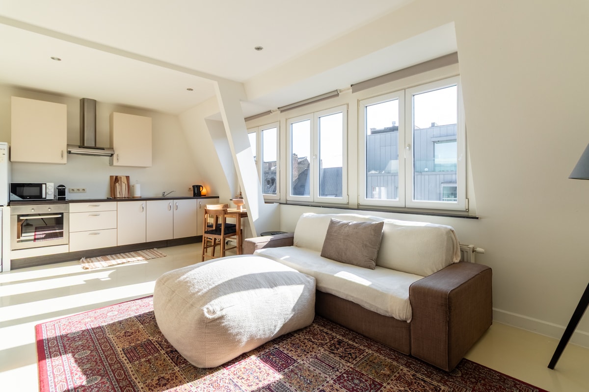A bright living area features large windows allowing natural light. A spacious sectional sofa is positioned on a patterned rug, with a small dining table and chairs visible in the background. Kitchen appliances are present, creating a functional open layout.