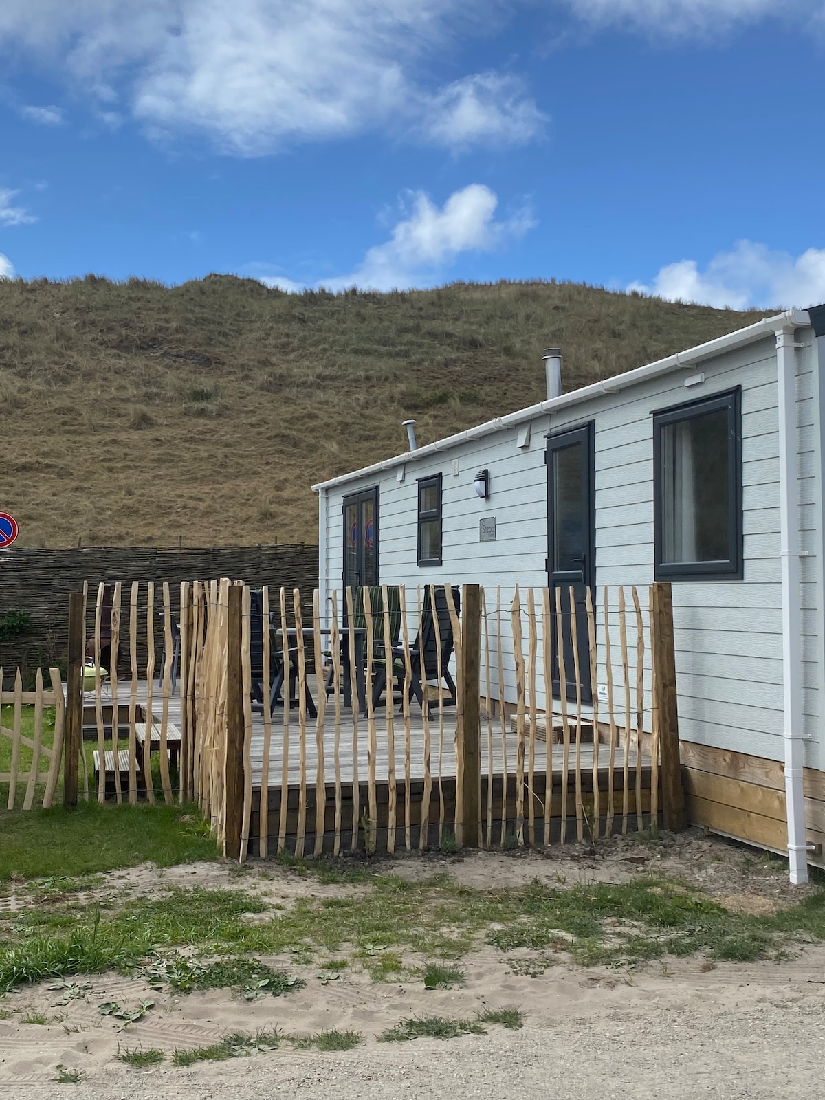 The exterior of a modern chalet is featured, showcasing a wooden deck surrounded by a bamboo fence. A grassy area is visible, with a hillside in the background and a clear blue sky above. Seating arrangements and potted plants can be seen on the deck.