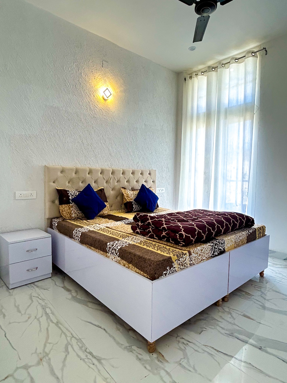 A bright bedroom features a spacious bed adorned with patterned bedding and decorative cushions. Natural light filters through sheer curtains, illuminating the room. Nightstands with drawers are placed on either side of the bed, and a ceiling fan adds to the comfort.