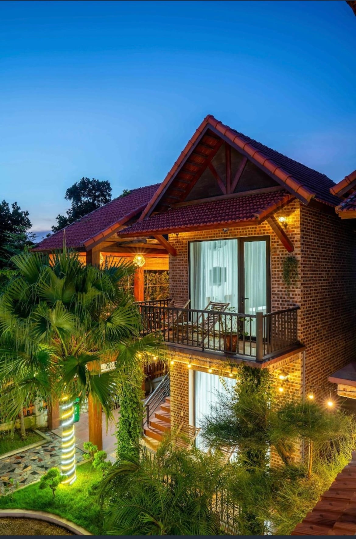 A charming brick structure is depicted, featuring a covered balcony adorned with soft curtains. Lush greenery surrounds the home, with palm trees adding to the inviting atmosphere. Warm lights illuminate the exterior, highlighting the architectural details against the twilight sky.