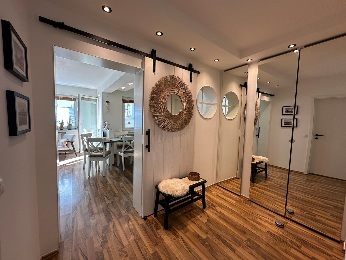 An inviting entryway is illuminated by recessed lighting, showcasing a cozy bench with a textured cushion. Circular mirrors and a decorative round mirror are mounted on the whitewashed wall, reflecting natural light. A wide, sliding barn door leads into a bright living area with a dining table.