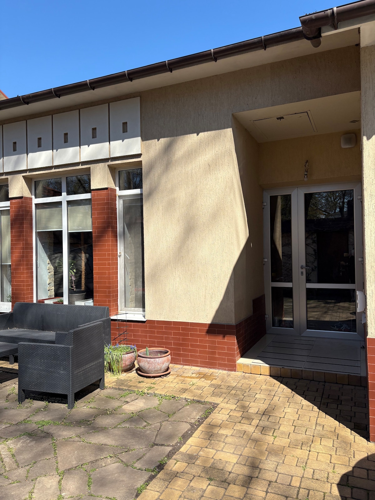 The exterior of the house is displayed, featuring large windows that invite natural light. A small entrance area is visible, flanked by decorative plants and a light-colored stone pathway. A shaded sitting area with dark outdoor furniture offers a space for relaxation.