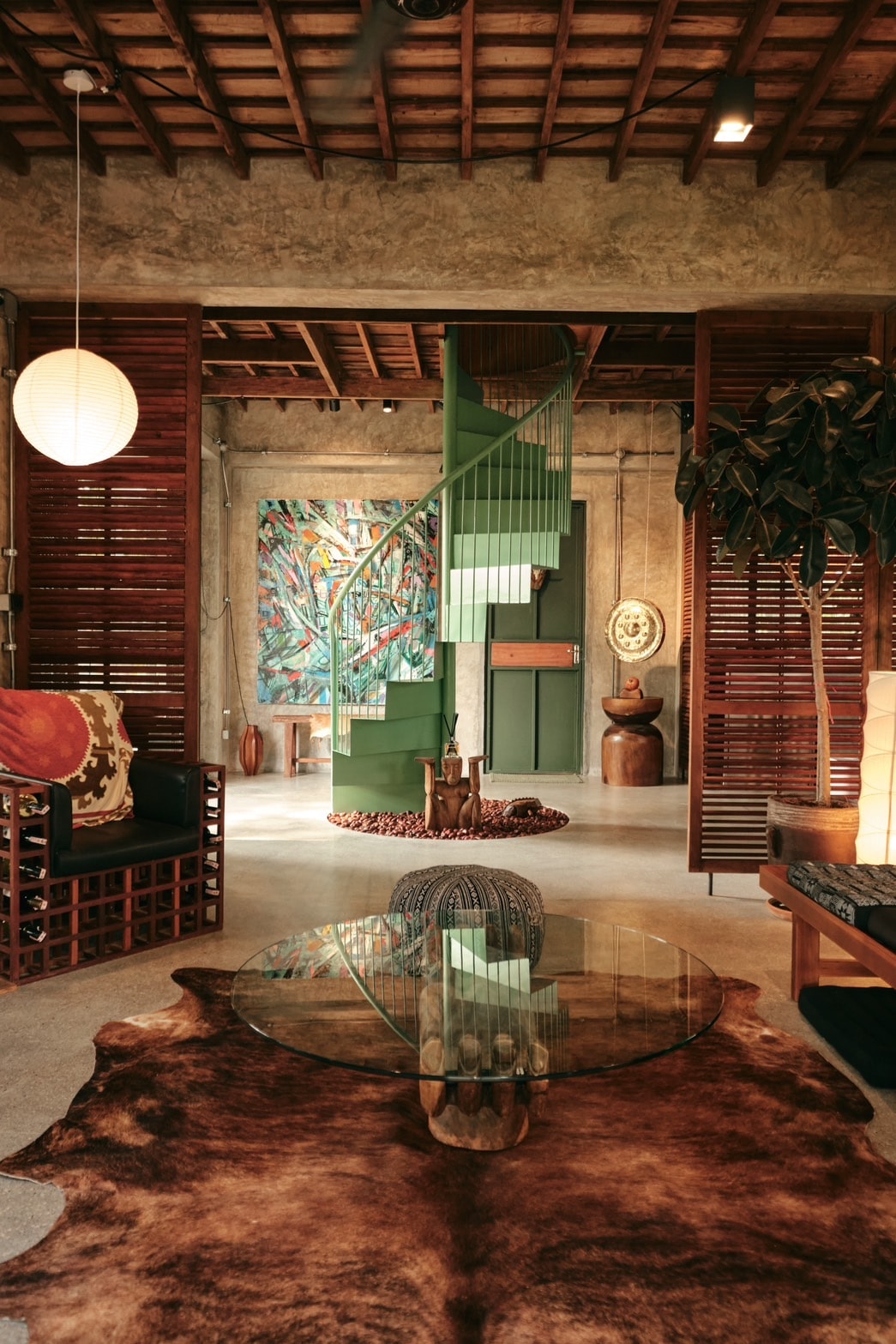An inviting living area features a circular glass coffee table atop a natural cowhide rug. A striking spiral staircase in a muted green leads upward, while vibrant wall art and a mix of wooden accents enhance the space's warmth. Soft lighting creates a relaxed ambiance.