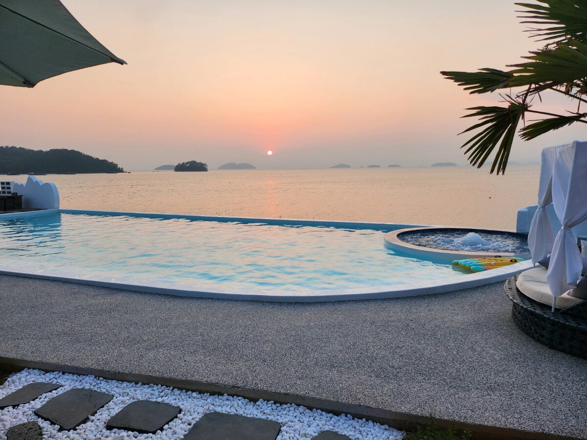 An outdoor infinity pool extends towards the horizon, reflecting the soft colors of the sunset over the ocean. A hot tub is positioned nearby, surrounded by pebbled walkways and lush greenery. Gentle ripples in the water create a peaceful ambiance.