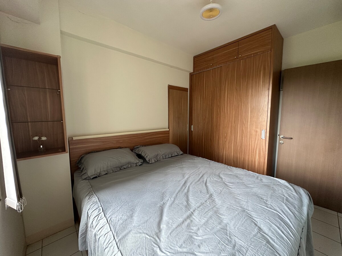A spacious sleeping area features a large bed with muted gray linens. Wood accents are visible on the wardrobe and furniture, complemented by natural lighting coming from a nearby window. The room includes neutral-colored walls and tiled flooring, creating a calm environment.