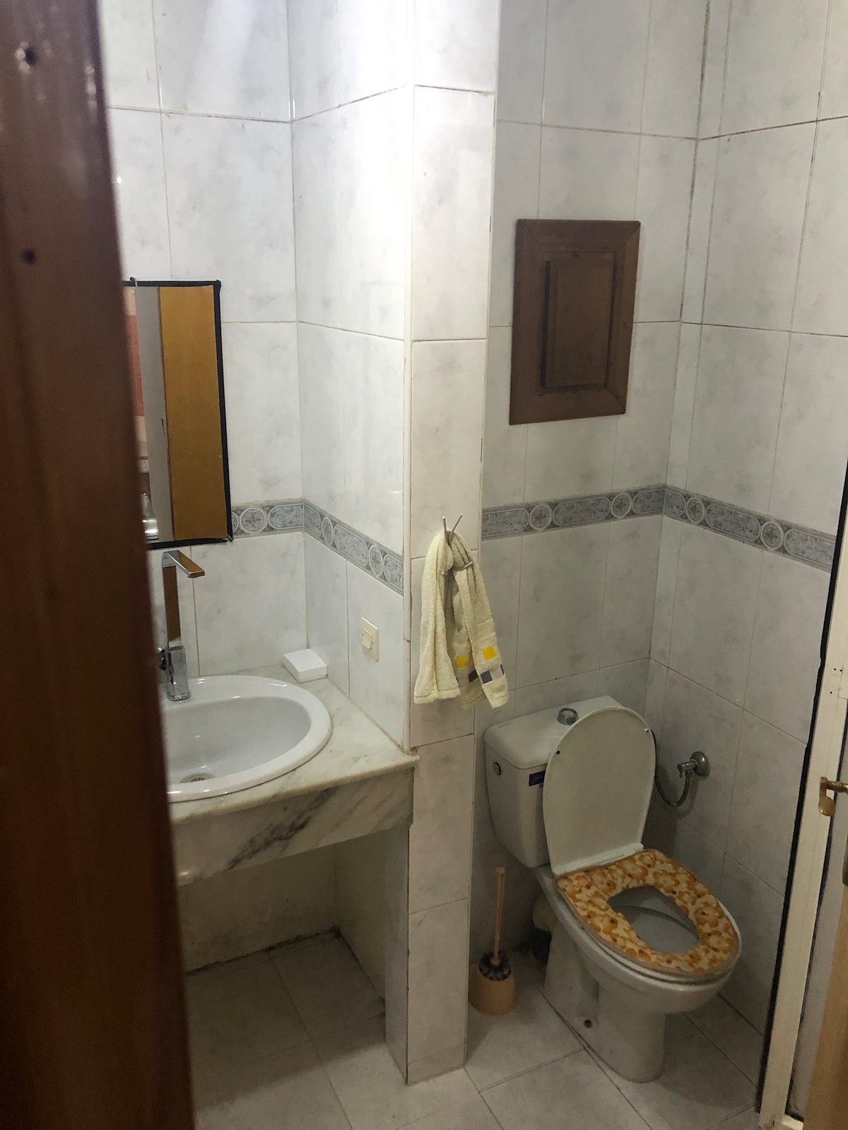 A compact bathroom space is featured, showcasing a white marble sink with a wall-mounted mirror above it. A neutral-colored toilet is positioned adjacent, complemented by a decorative toilet seat cover. White tiled walls create a clean and bright atmosphere.