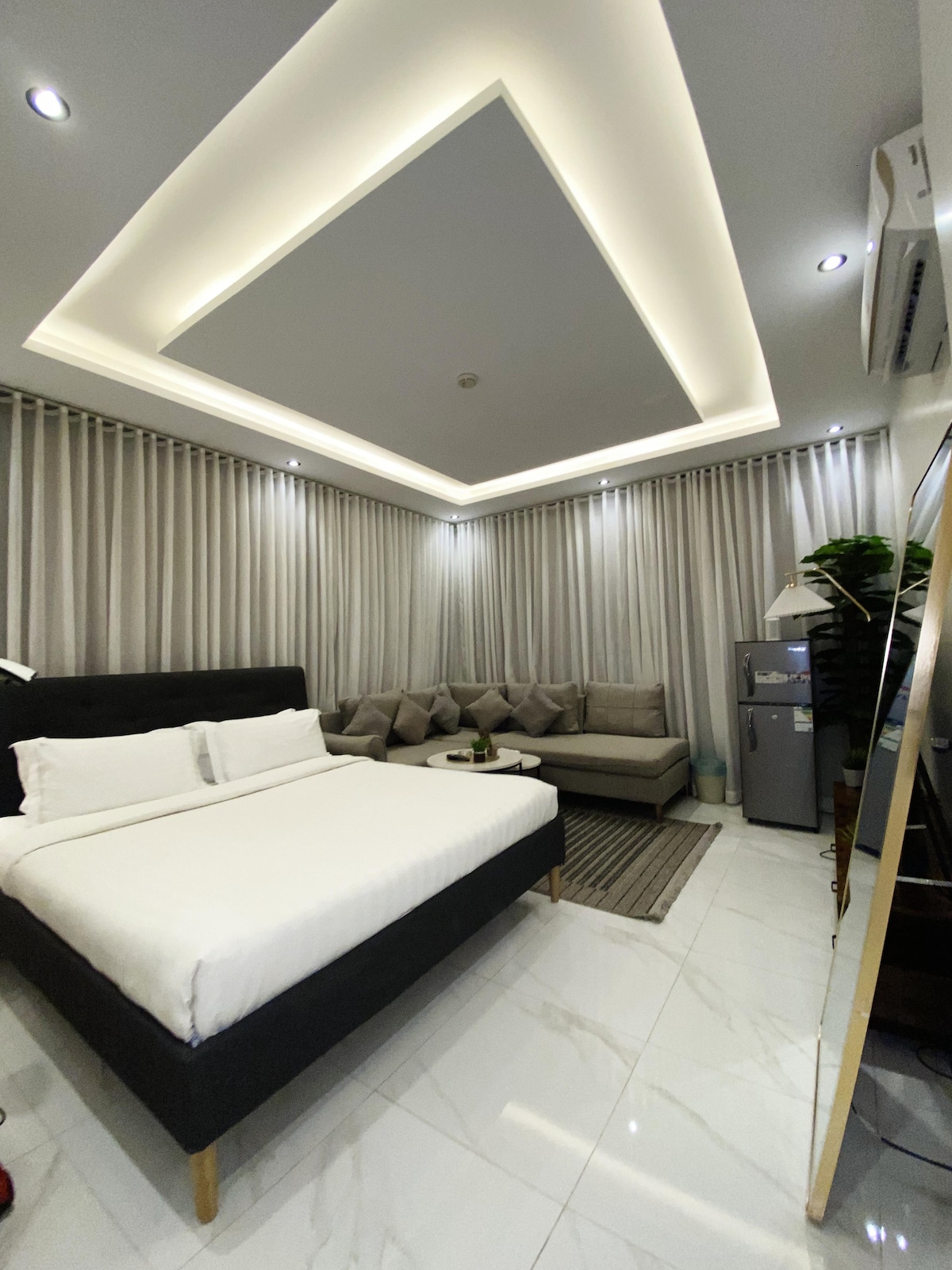 A spacious room features a comfortable bed with crisp linens and a well-arranged seating area with plush cushions. Large curtains frame the windows, while modern lighting casts a soft glow across the room's sleek tiled floor. A mini-fridge is discreetly positioned nearby.