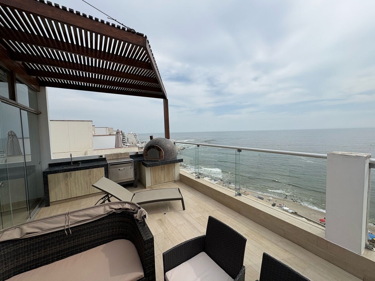 An expansive terrace offers a view of the ocean and coastline. Comfortable outdoor seating surrounds a stone oven, and a grill area is visible. A partially shaded area is provided by wooden trellising. The horizon shows a subtle blend of sea and sky.