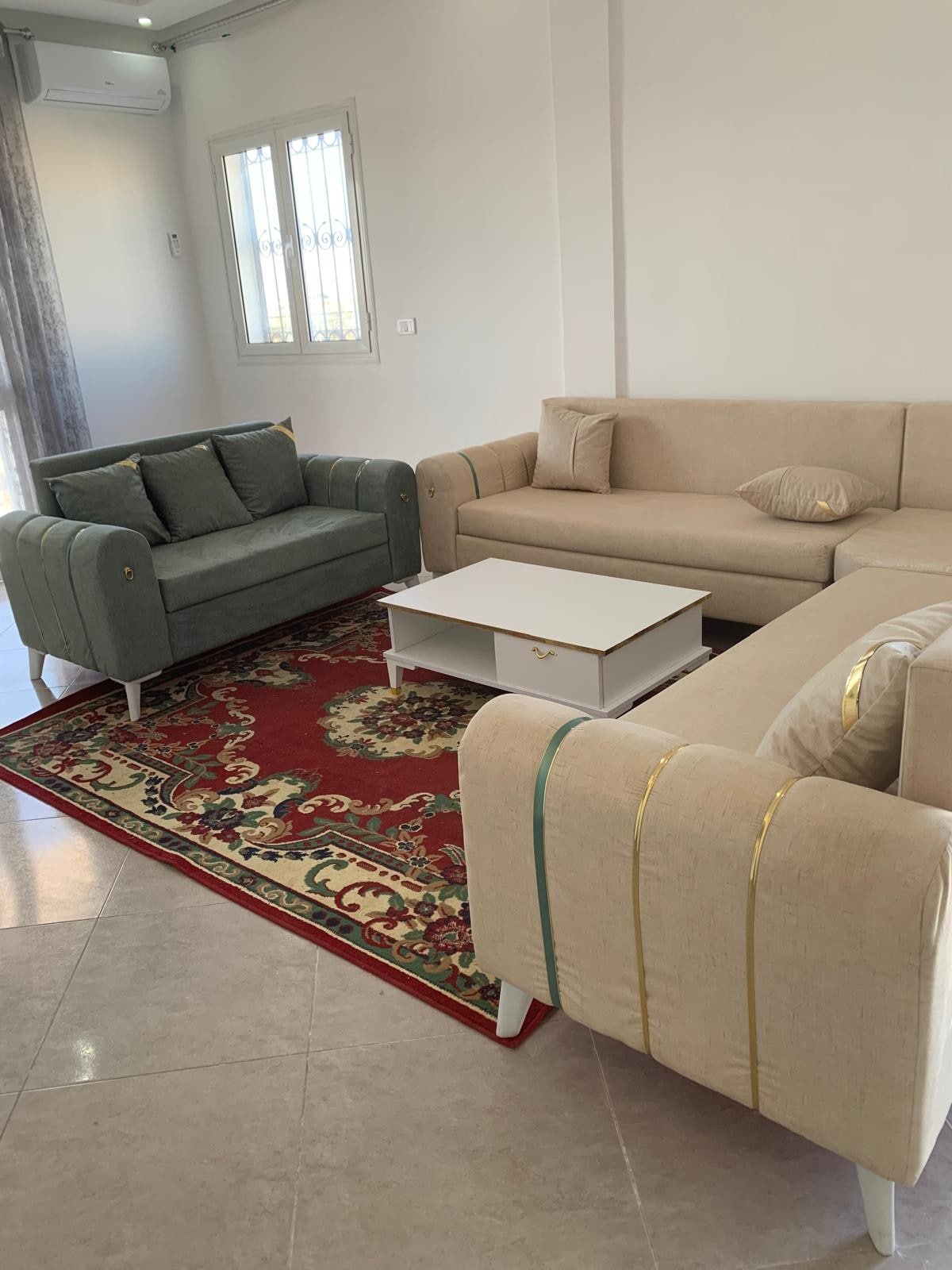 A cozy living area showcases a combination of light-colored sofas with green accents arranged around a white coffee table. A patterned area rug adds a touch of warmth, while large windows invite natural light, creating an open and welcoming space.