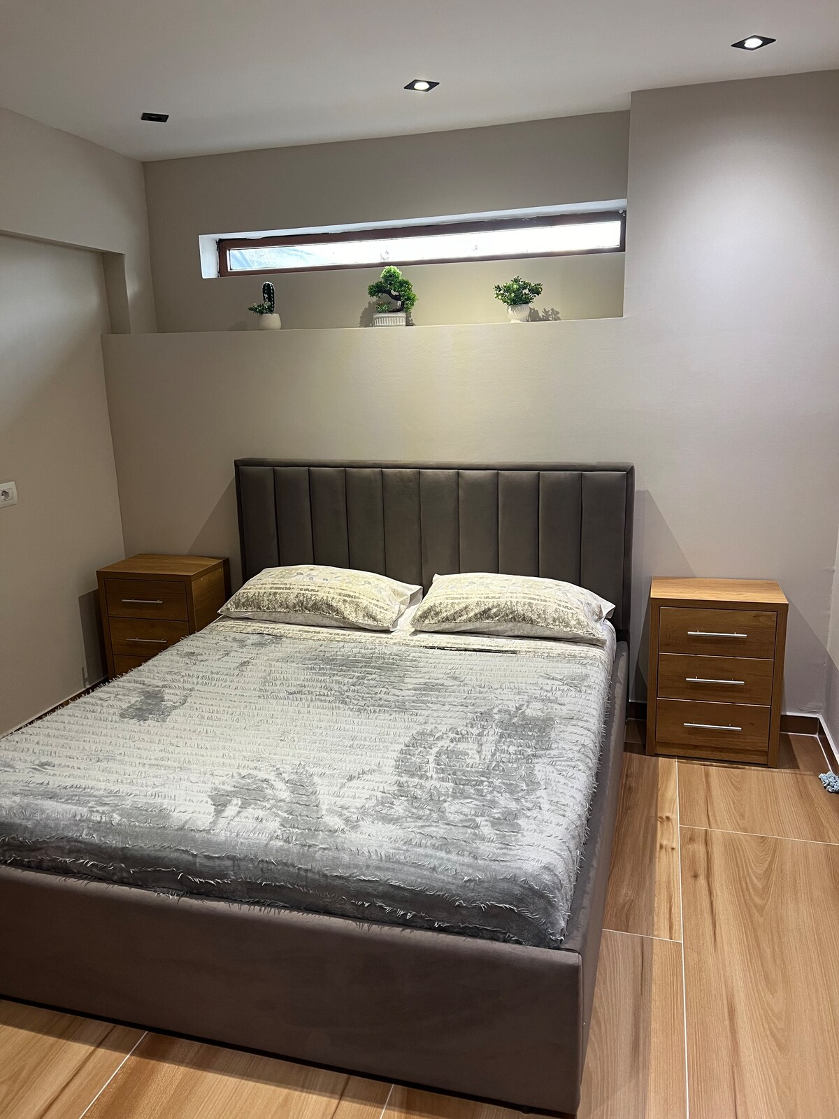 A cozy bedroom features a neatly made, spacious bed with soft grey bed linens. Two wooden nightstands are positioned on either side of the bed. Natural light filters through a small window, illuminating minimalistic decorations placed on the ledge above.