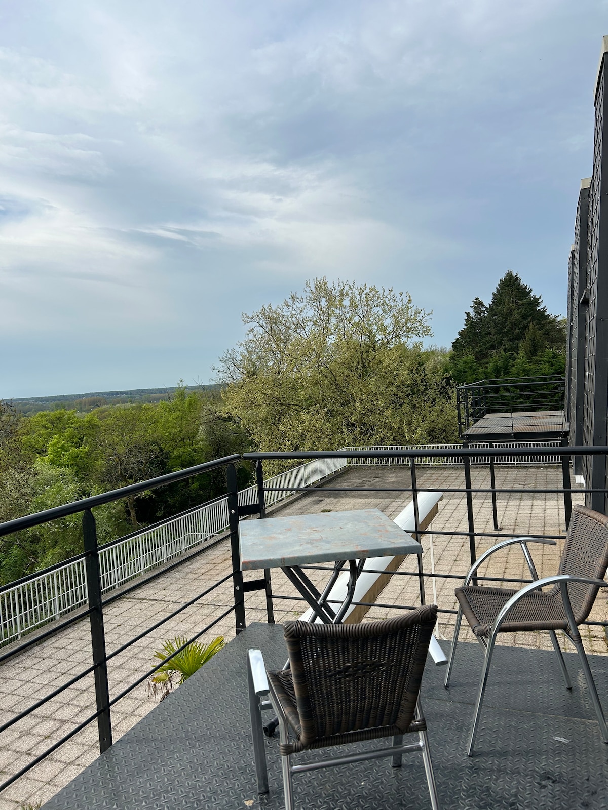 A balcony terrace is visible, featuring a small table and three chairs. Lush greenery is observed in the distance, with a view extending beyond the boundary. The surrounding area includes paved flooring and a railing, contributing to an open and airy atmosphere.