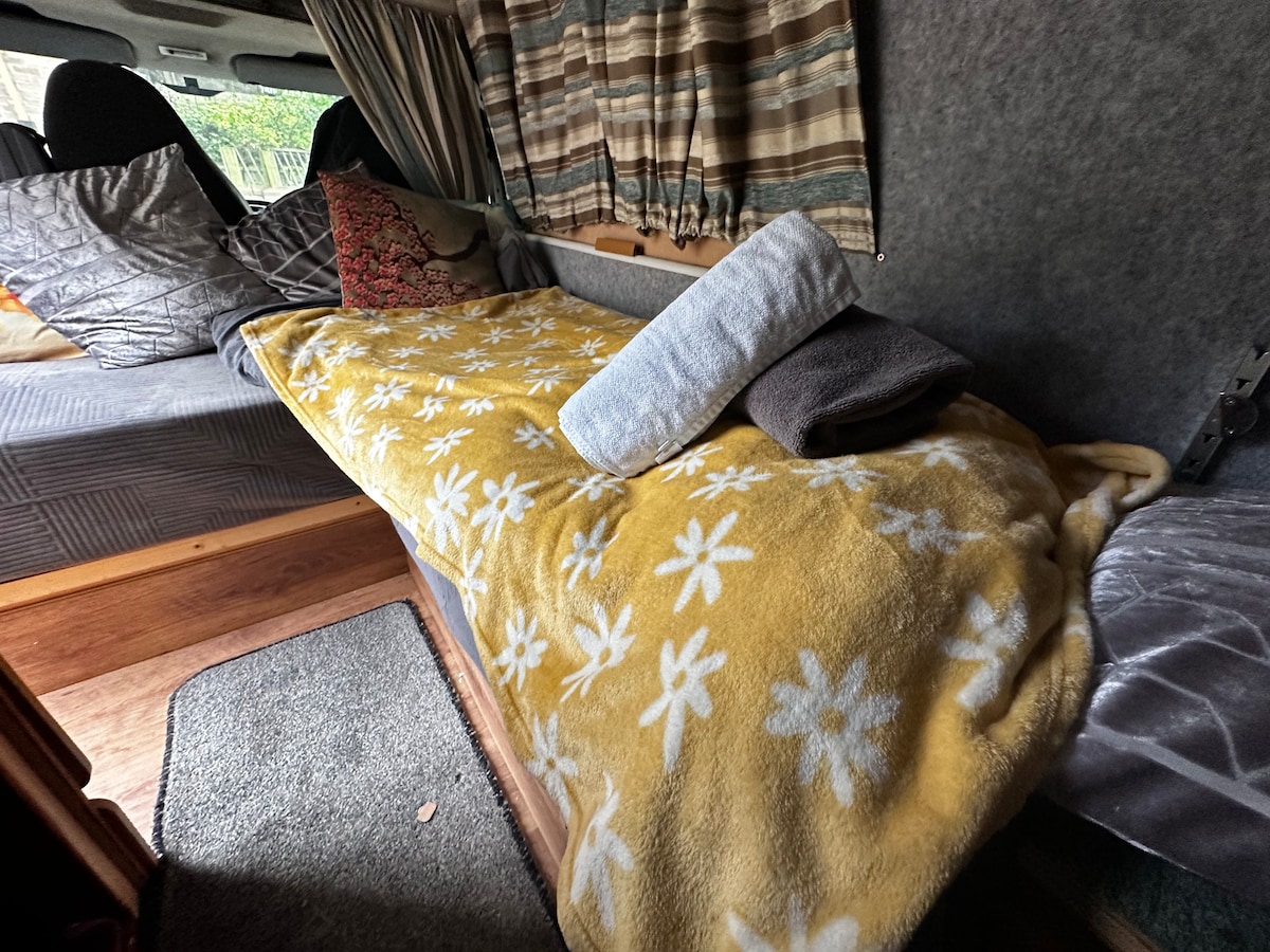 A comfortable sleeping area is presented with a soft, floral-patterned blanket draped over the bed. Grey and beige towels are neatly arranged at the foot of the bed, and patterned cushions add a cozy touch to the space. Natural light filters through the curtains.