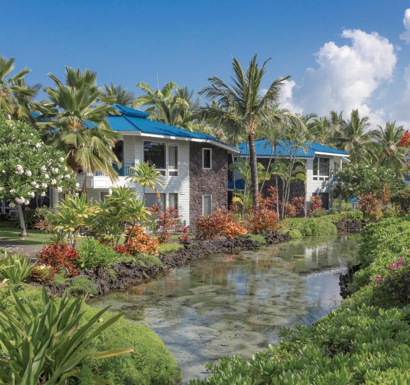 Lush tropical landscaping surrounds the resort, featuring various flowering plants and palm trees. A tranquil water feature flows nearby, reflecting the clear blue sky and vibrant greenery, while the two-story lanai-styled buildings provide a serene setting for relaxation.