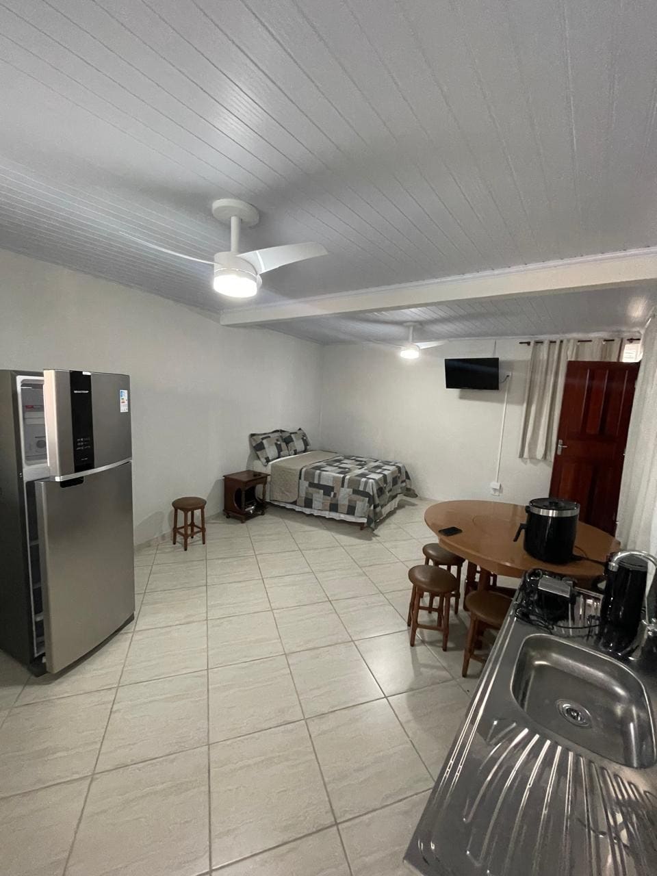 A compact living area is visible, featuring a spacious bed with subtle patterned bedding, accompanied by two small stools and a round dining table. A modern refrigerator stands against one wall, while a sink area and a television are positioned nearby. Bright ceiling lights illuminate the space.