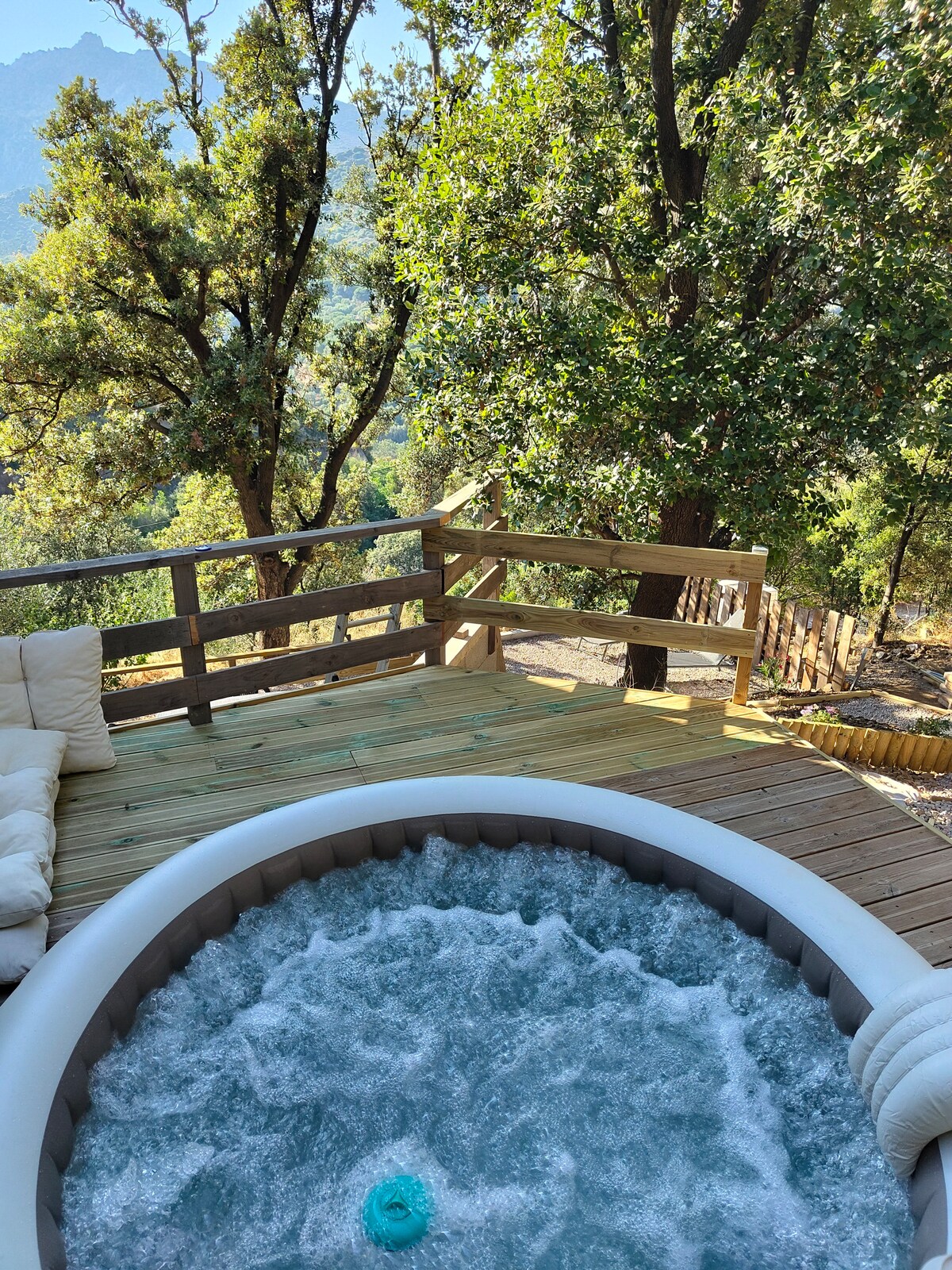 A jacuzzi is situated on a wooden deck, surrounded by lush greenery and trees. The clear water creates gentle bubbles, and the relaxing outdoor environment offers views of the mountainous terrain in the distance.