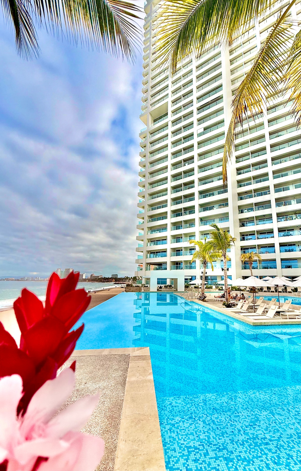 A tranquil pool area is framed by swaying palm trees and colorful tropical flowers. The modern building rises alongside the stunning water, with lounge chairs placed around the pool's edge. The clear blue water reflects the sun and surrounding architecture.