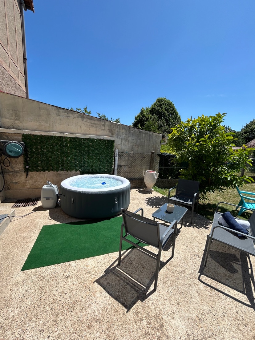 An outdoor space features a circular hot tub surrounded by a green lawn and seating arrangements. Two metal chairs are positioned on a light stone patio, along with a small table. Sunlight brightens the area, enhancing the inviting atmosphere of the garden.