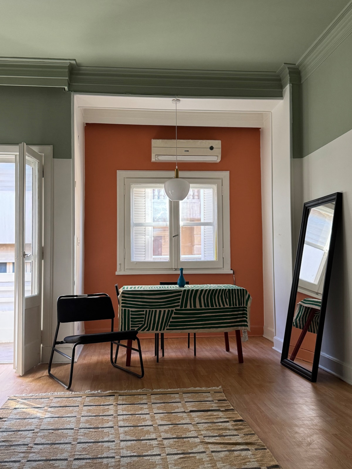 A bright room displays a small table draped in a green-striped cloth, accompanied by a black chair. A wall-mounted air conditioner is positioned above a window, framed by white shutters. Light reflects off a large mirror, enhancing the sense of space.