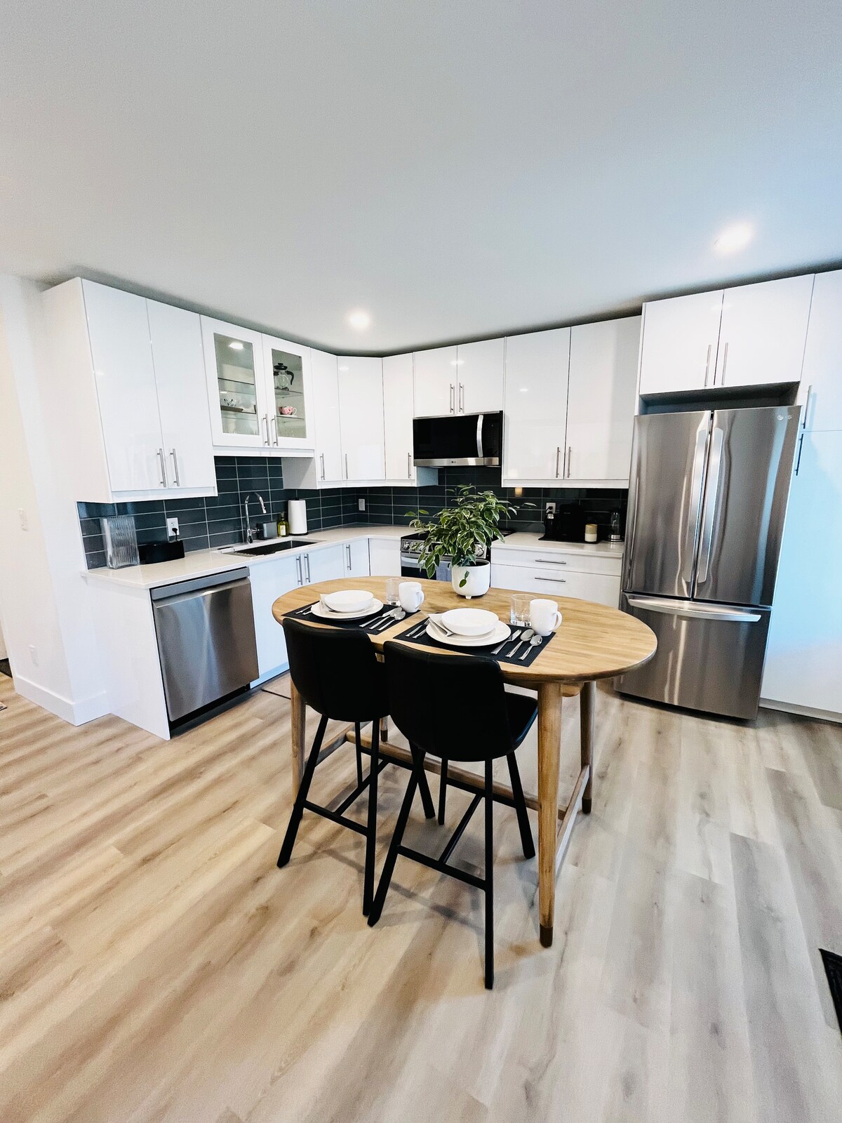 A modern kitchen features sleek white cabinetry paired with dark countertops, creating a clean aesthetic. A round wooden table is set for two, with stylish tableware and a small green plant adding a touch of freshness. Stainless steel appliances and ample natural light complete the inviting space.