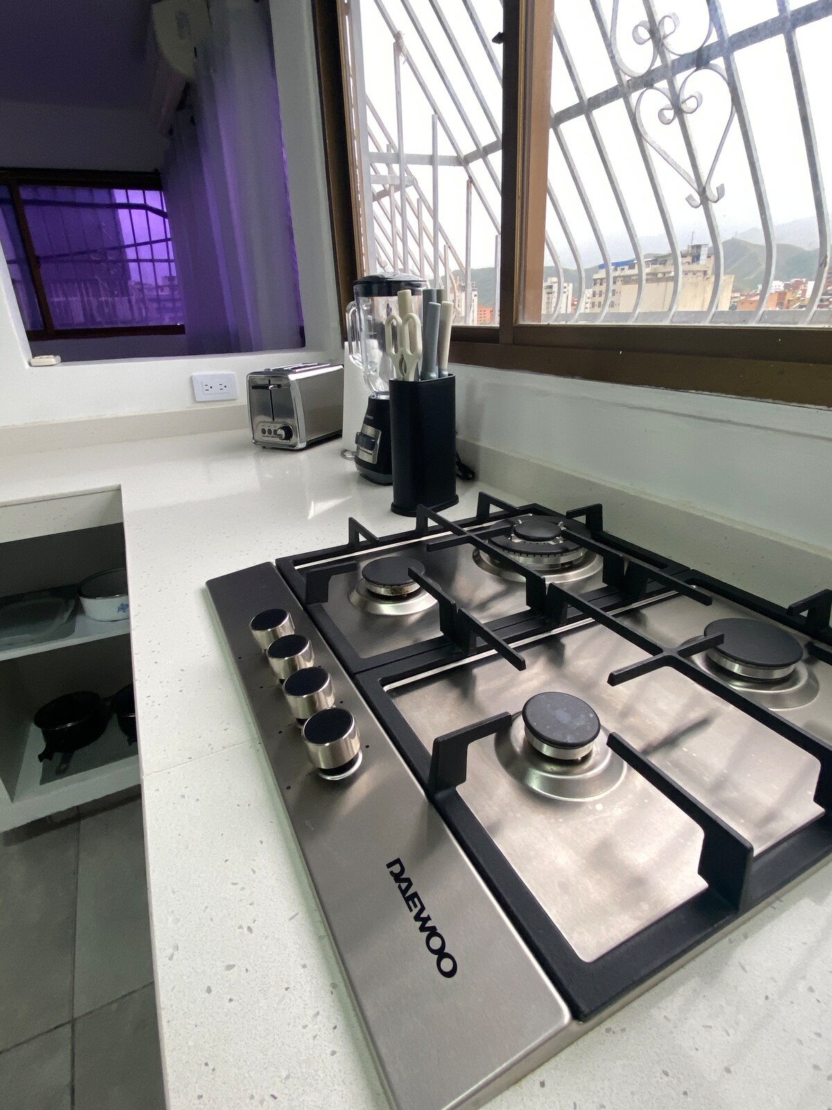 A modern kitchen countertop is highlighted, featuring a stainless steel gas stove with five burners. A blender and toaster are situated nearby, alongside a knife block. Natural light enters through a window adorned with white curtains, revealing distant mountain views.